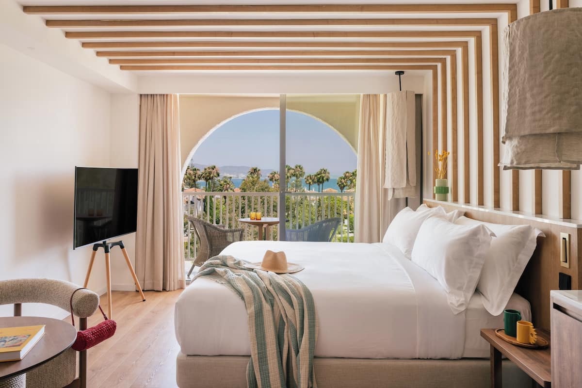 A hotel room with a large bed, a TV, a balcony with a view of the sea, and a dining area with a table and chairs.