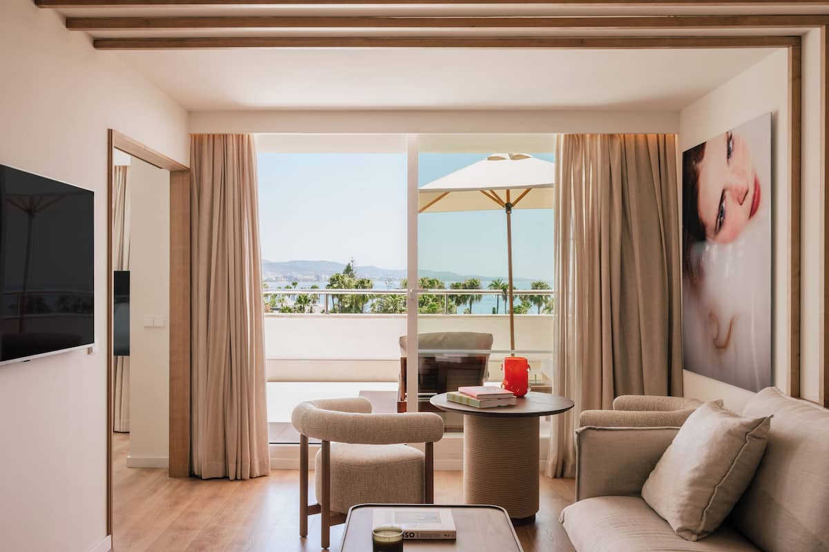A modern hotel room with a balcony, a sofa, a chair, a table with a book and a red candle, and a TV mounted on the wall.