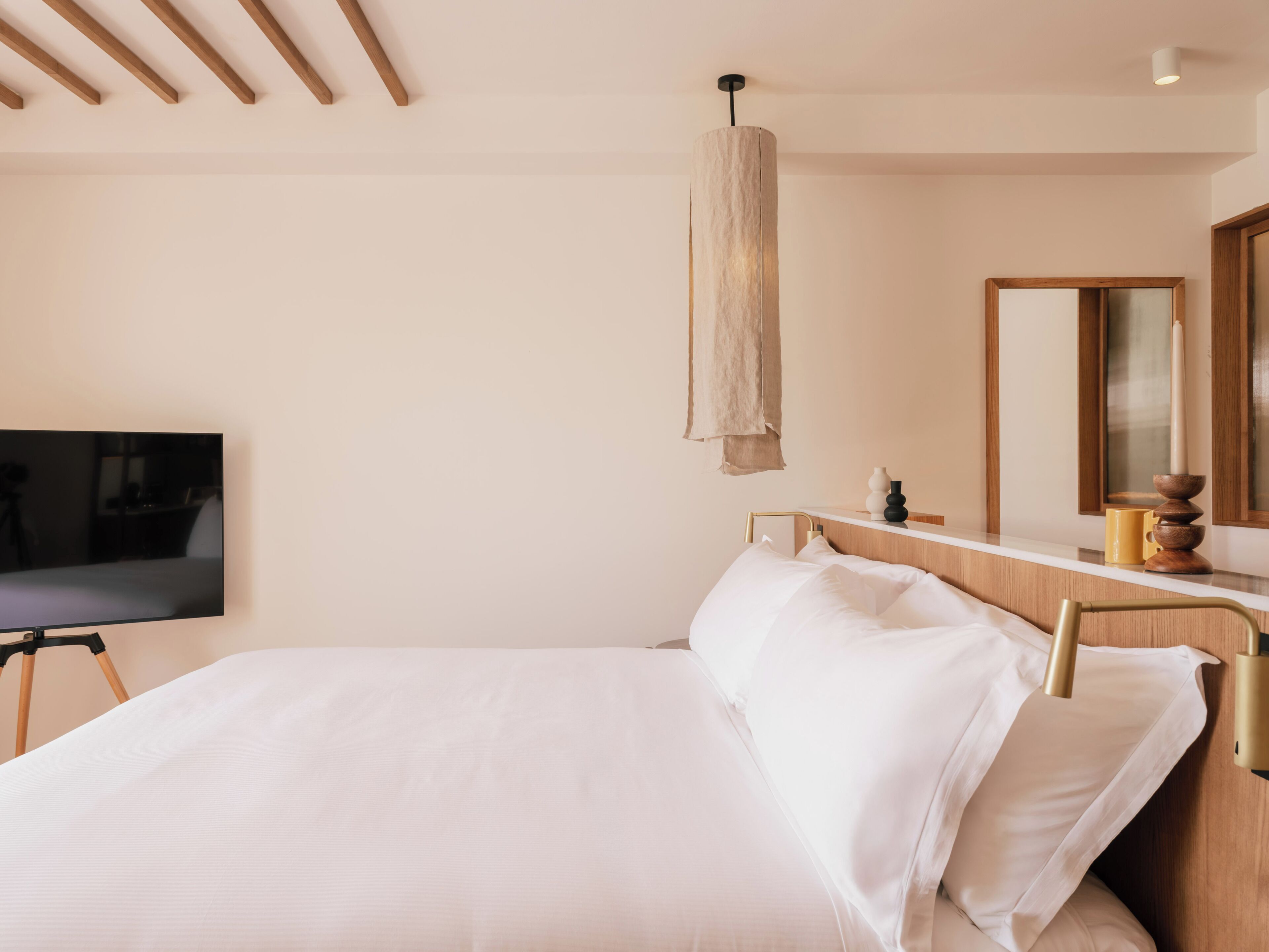 A modern hotel room with a bed, a flat-screen TV, a wooden headboard, a bedside table with a vase and a candle, and a mirror.