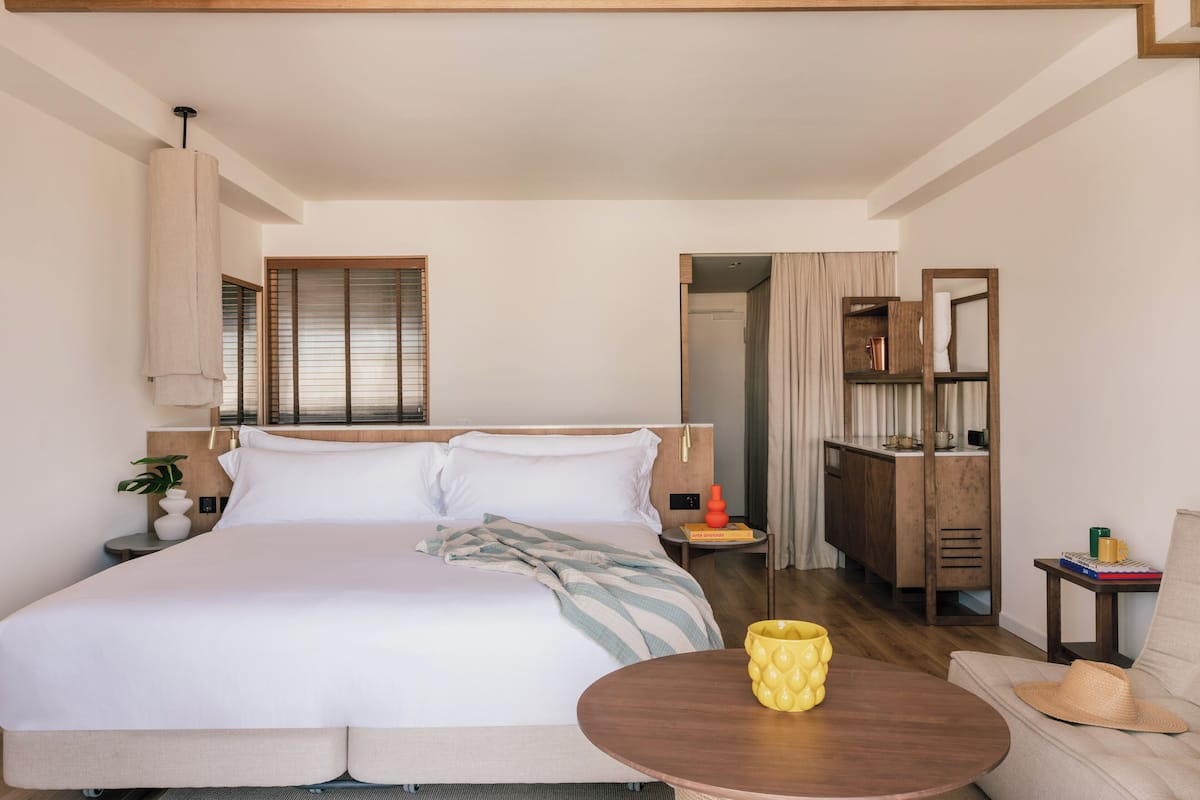 A hotel room with a large bed, a bedside table, a small round table, a chair, a wardrobe, and a window with blinds.