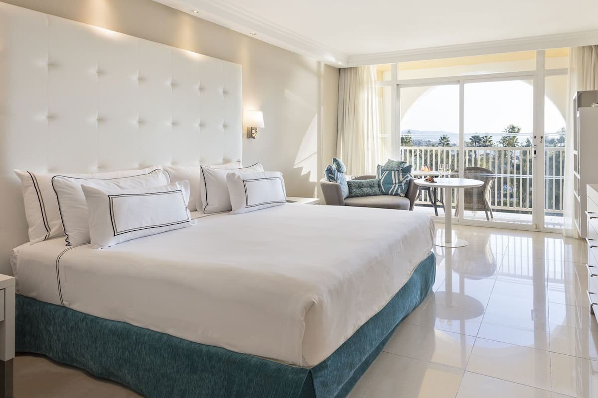 A hotel room with a large bed, a seating area with a table and chairs, and a balcony with a view.