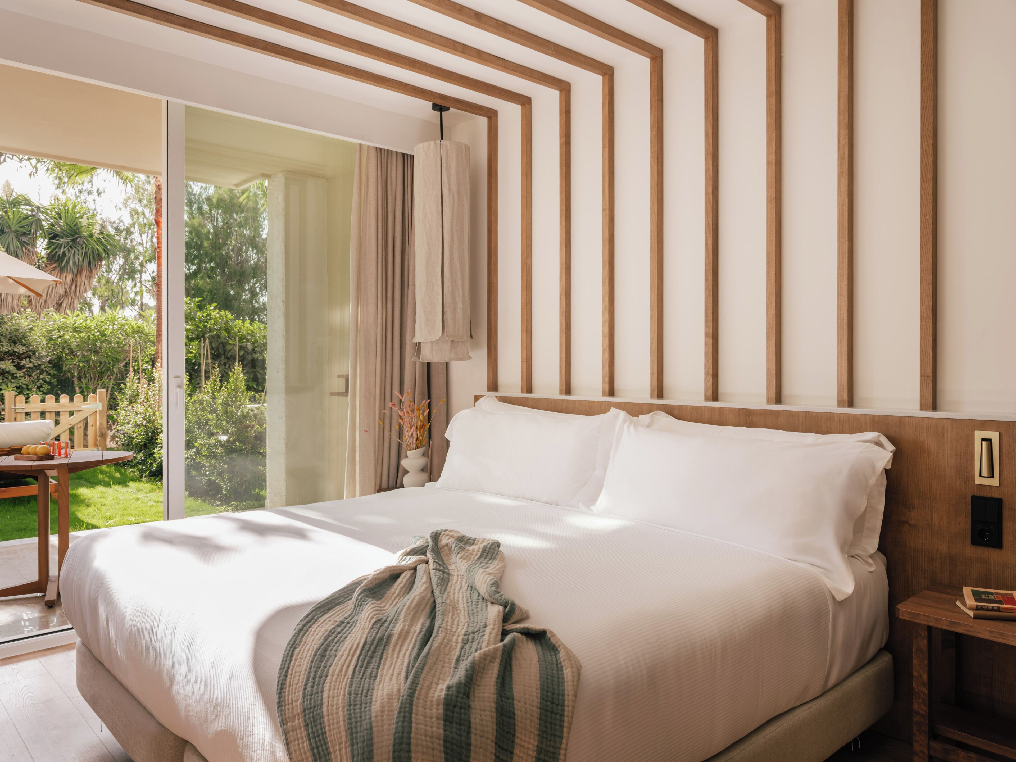 Un dormitorio con una cama grande, cabecera de madera, una mesita de noche con un libro y vistas a un jardín a través de una puerta de cristal.