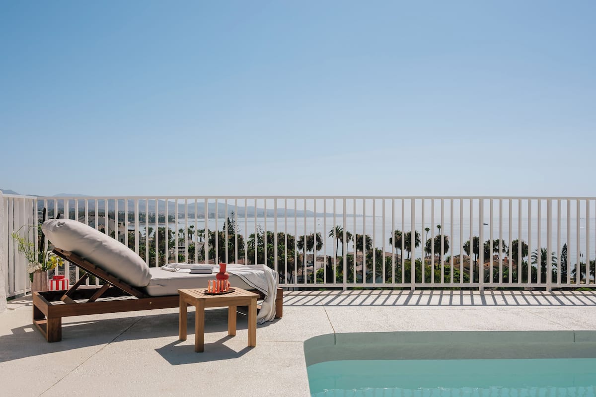A sun lounger with a cushion, a small table with a vase, and a poolside view of a coastal area with palm trees and buildings.