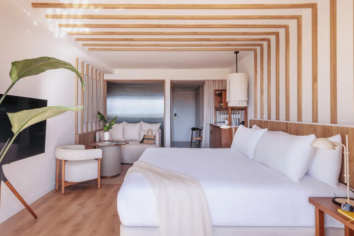 A modern hotel room with a large bed, a sitting area with a chair and table, a large plant, and a view of the sea through an open door.