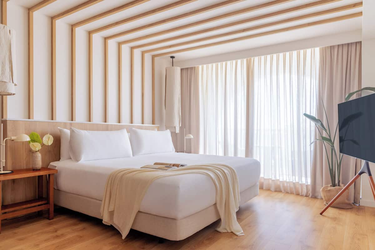 A modern hotel room with a large bed, wooden floor, and a view of the city through large windows.