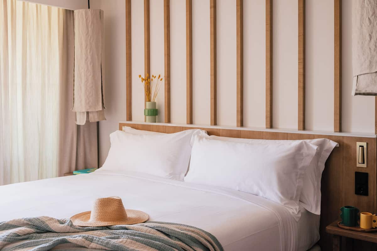 A neatly made bed with white linens, a woven straw hat, and a striped blanket.