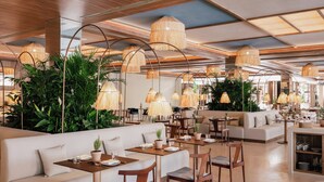 Un restaurant au décor moderne, avec des panneaux de bois au plafond, des lampes en rotin suspendues et des plantes vertes. On y trouve plusieurs espaces de restauration avec des tables et des chaises.