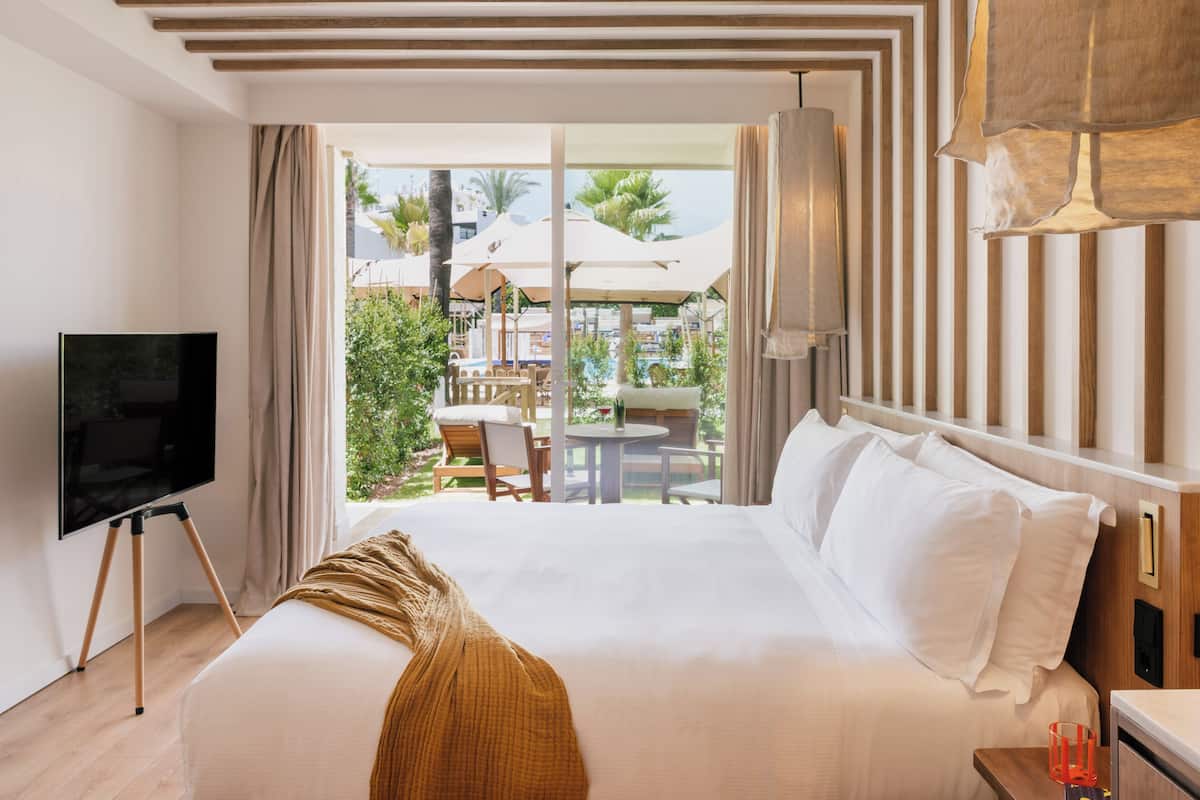 A hotel room with a large bed, a TV on a stand, and a view of the outdoor area with umbrellas and palm trees.