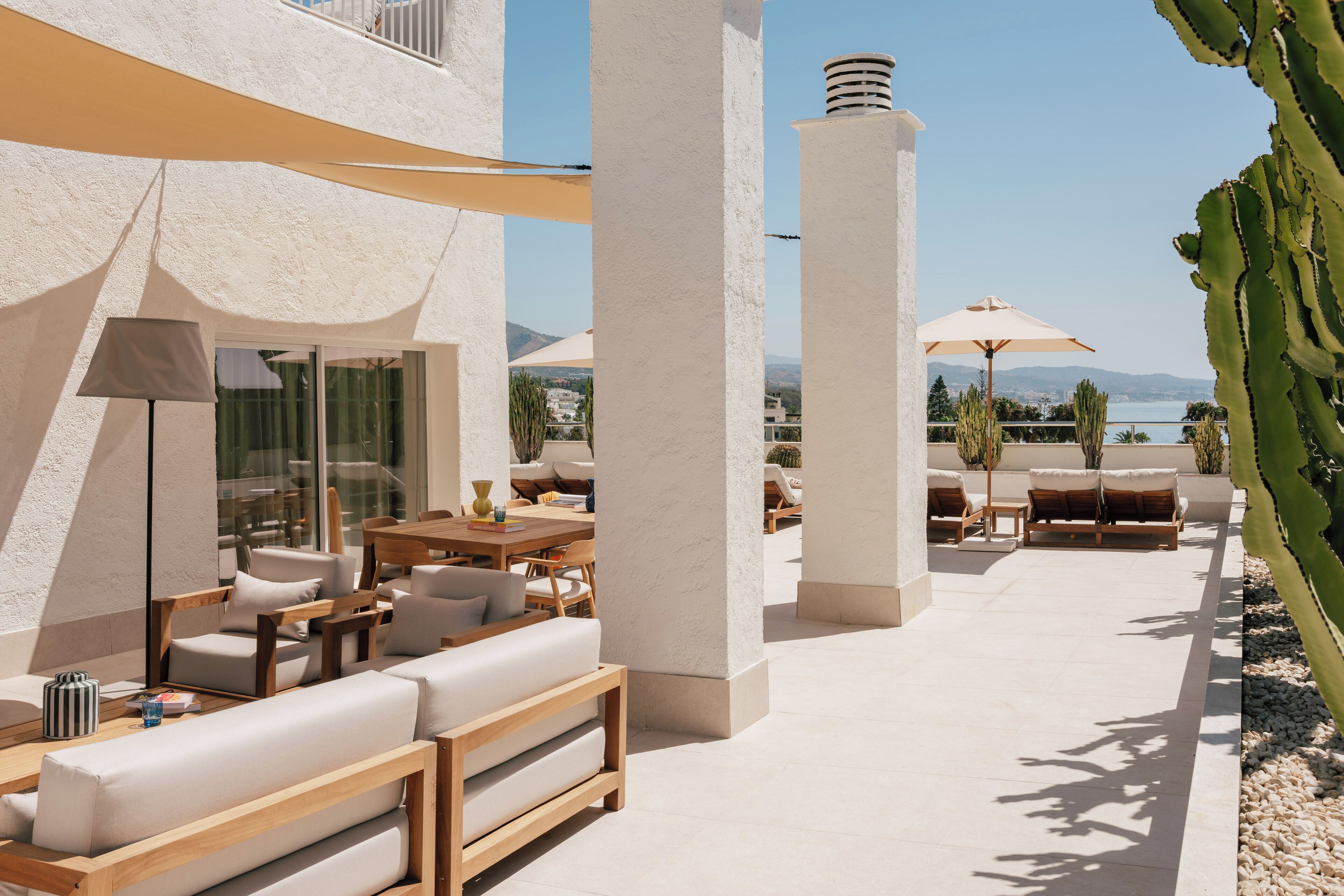 An outdoor seating area with white sofas, wooden tables, and umbrellas, overlooking a body of water and mountains.