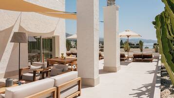 An outdoor seating area with white sofas, wooden tables, and umbrellas, overlooking a body of water and mountains.