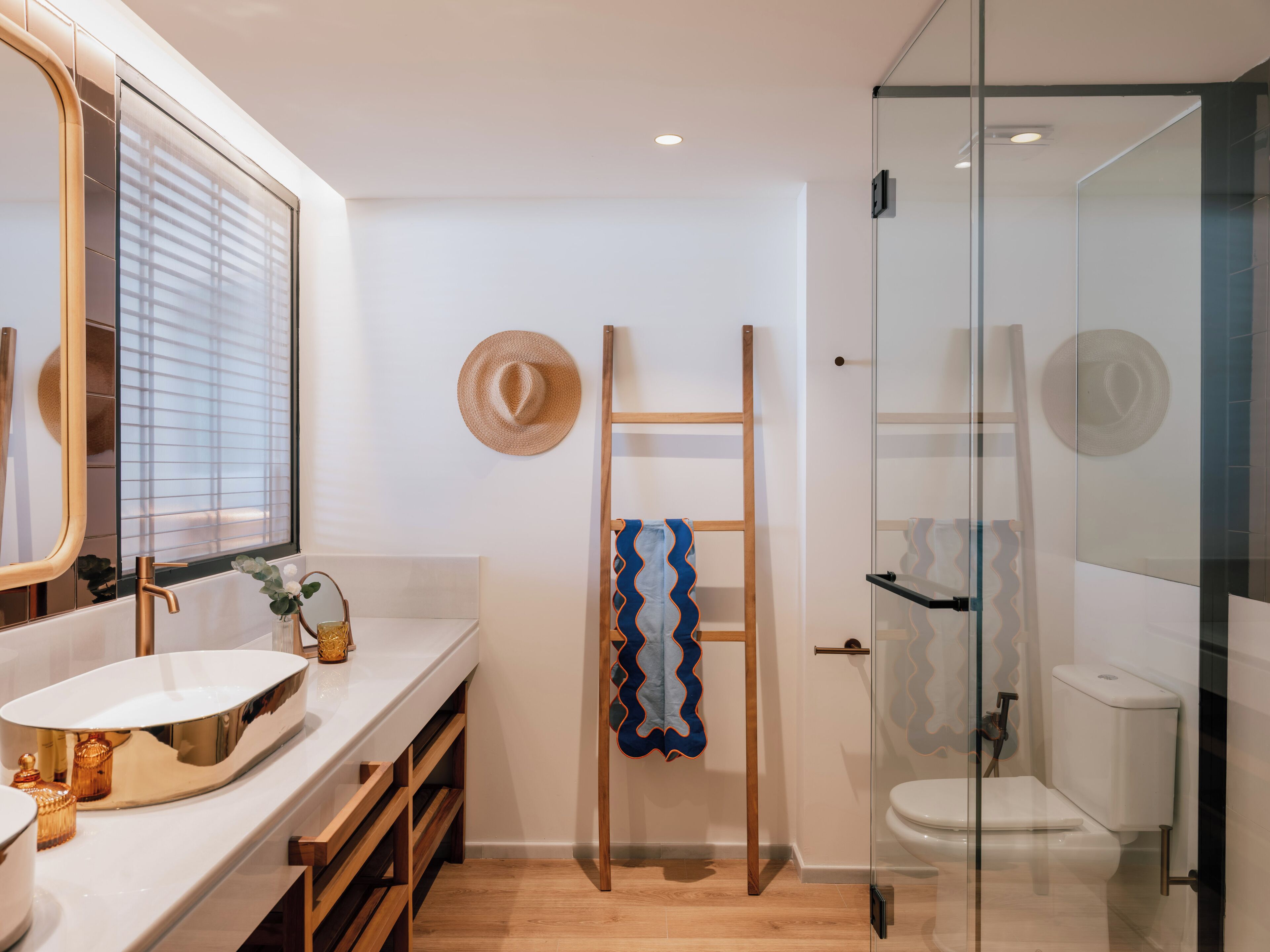 Un baño moderno con un espejo grande, tocador blanco, repisas de madera, un perchero con un sombrero y cabina de ducha de vidrio.