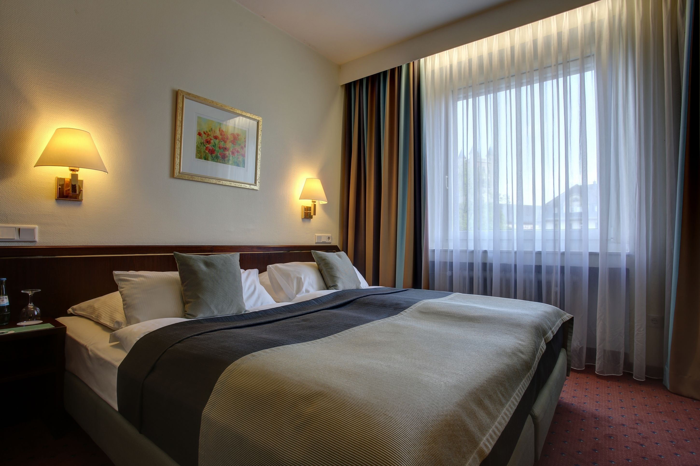 Habitación de hotel con una cama grande, dos lámparas de noche, un cuadro enmarcado en la pared y una ventana con cortinas traslúcidas.