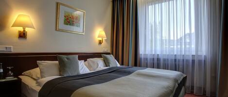Habitación de hotel con una cama grande, dos lámparas de noche, un cuadro enmarcado en la pared y una ventana con cortinas traslúcidas.