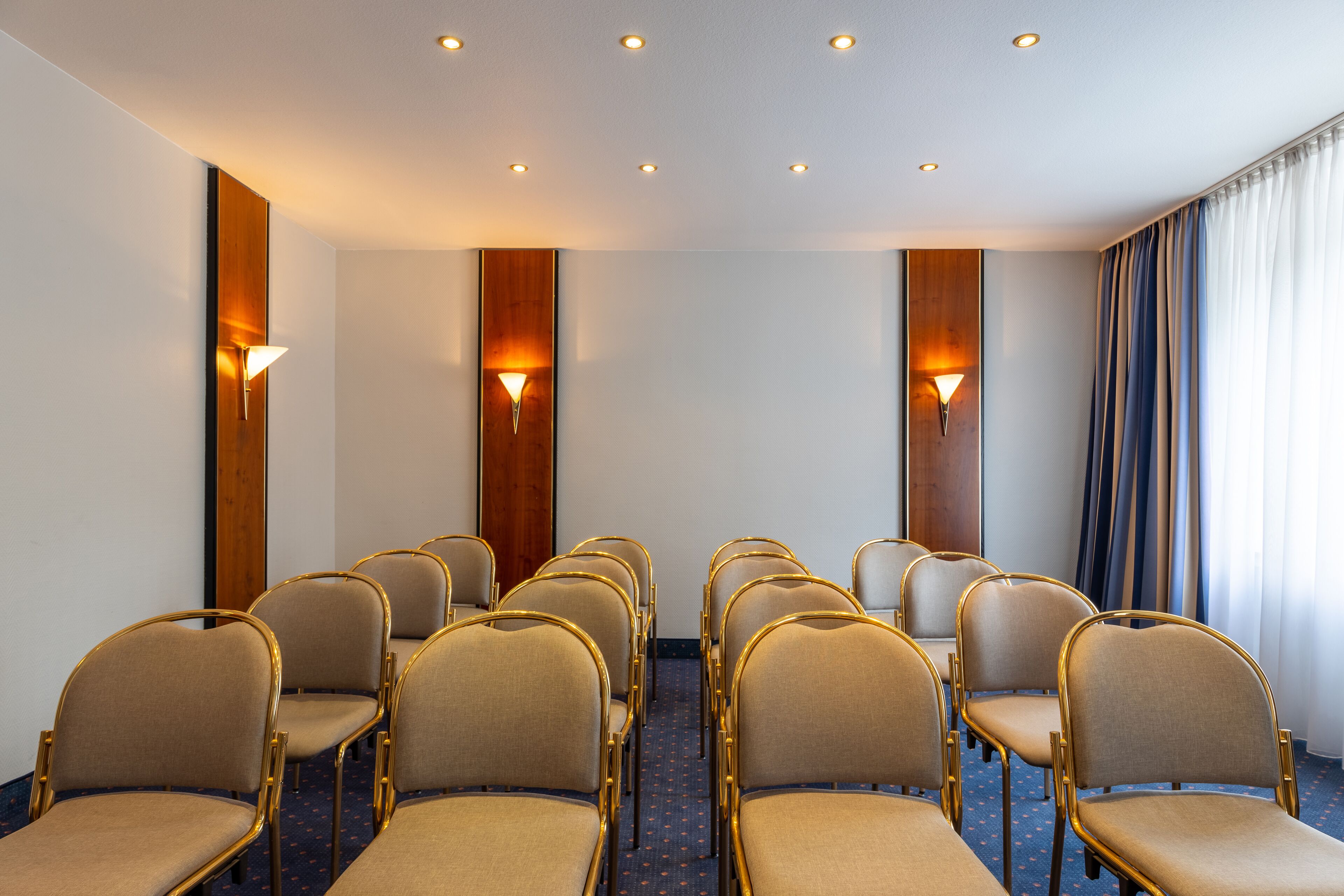 A conference room with rows of beige chairs and wooden paneling, equipped with wall-mounted lamps.