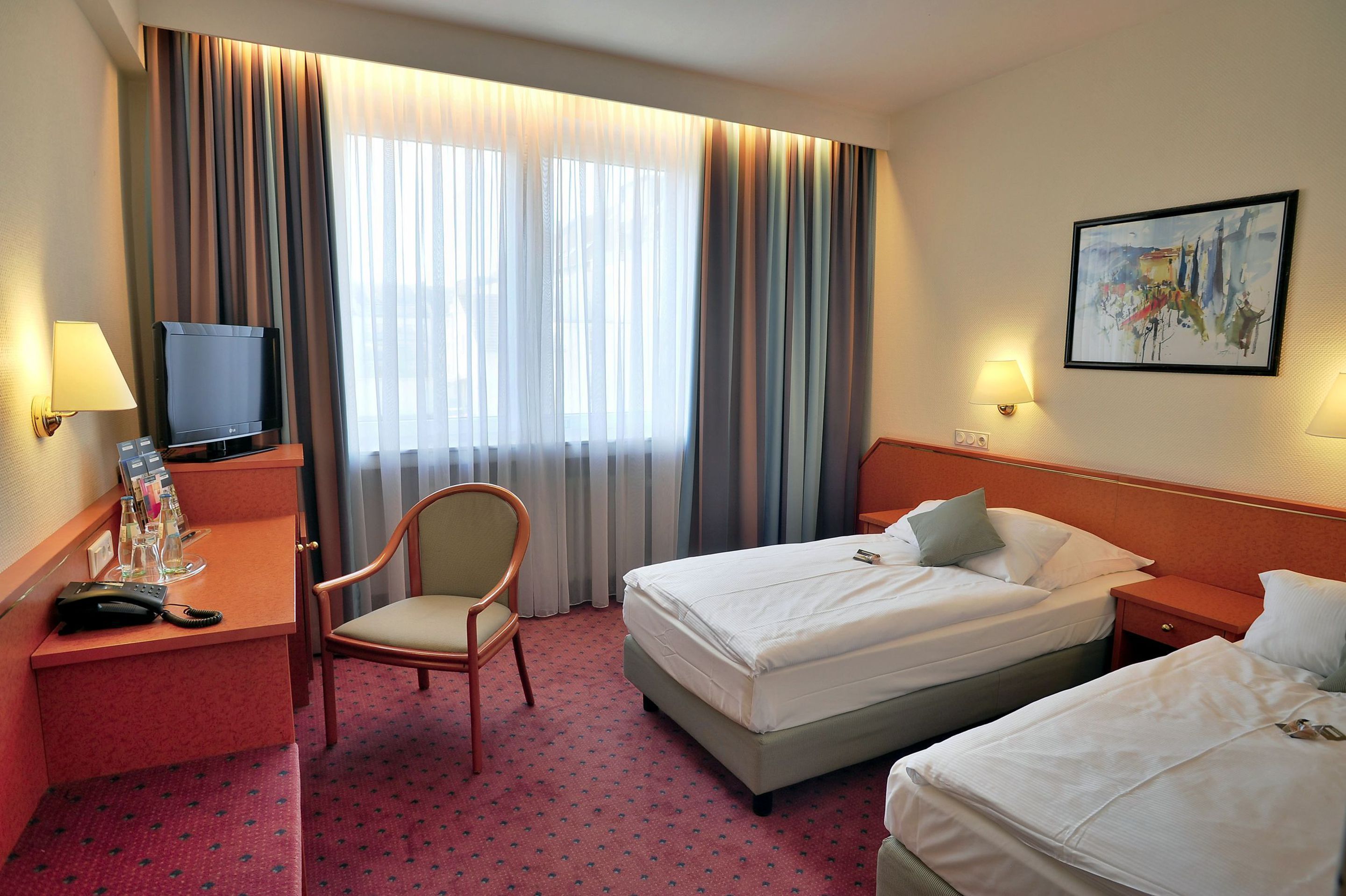 A hotel room with two beds, a desk with a TV, a chair, and a painting on the wall.
