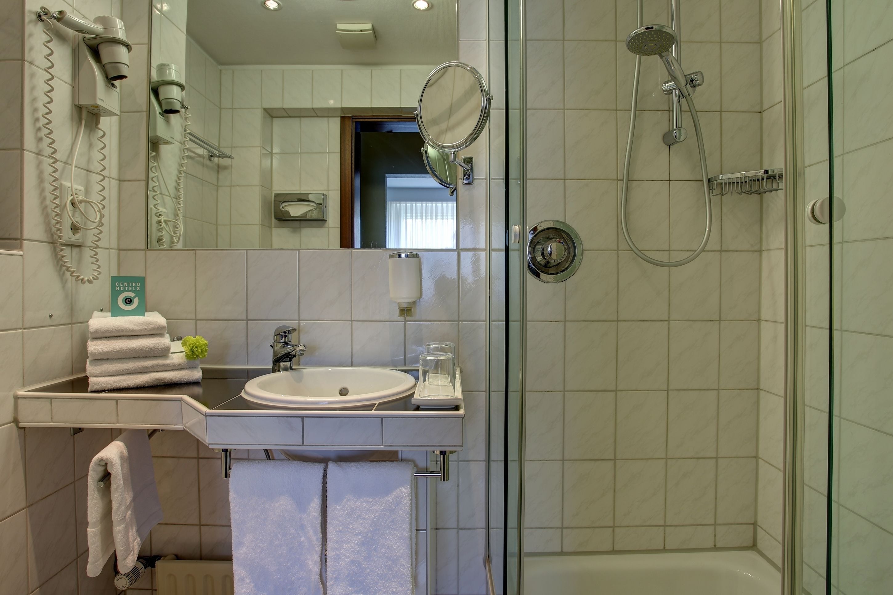 Une salle de bain moderne équipée d’un lavabo, d’un miroir, d’un sèche-cheveux et d’une douche.