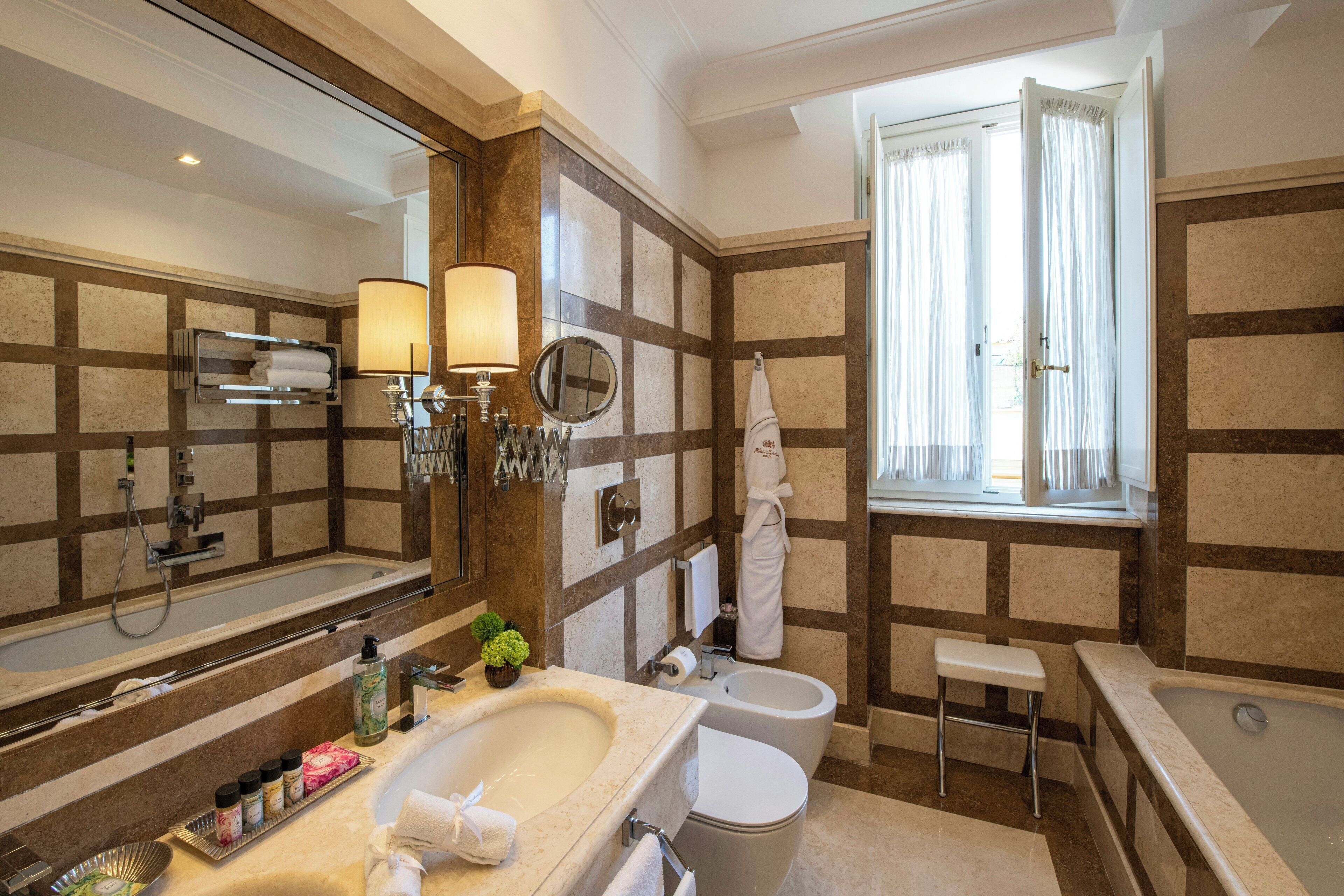 Un baño con un espejo grande, una bañera, un lavamanos y un inodoro. Hay una ventana con cortinas, un perchero para toallas y una planta pequeña en la encimera.