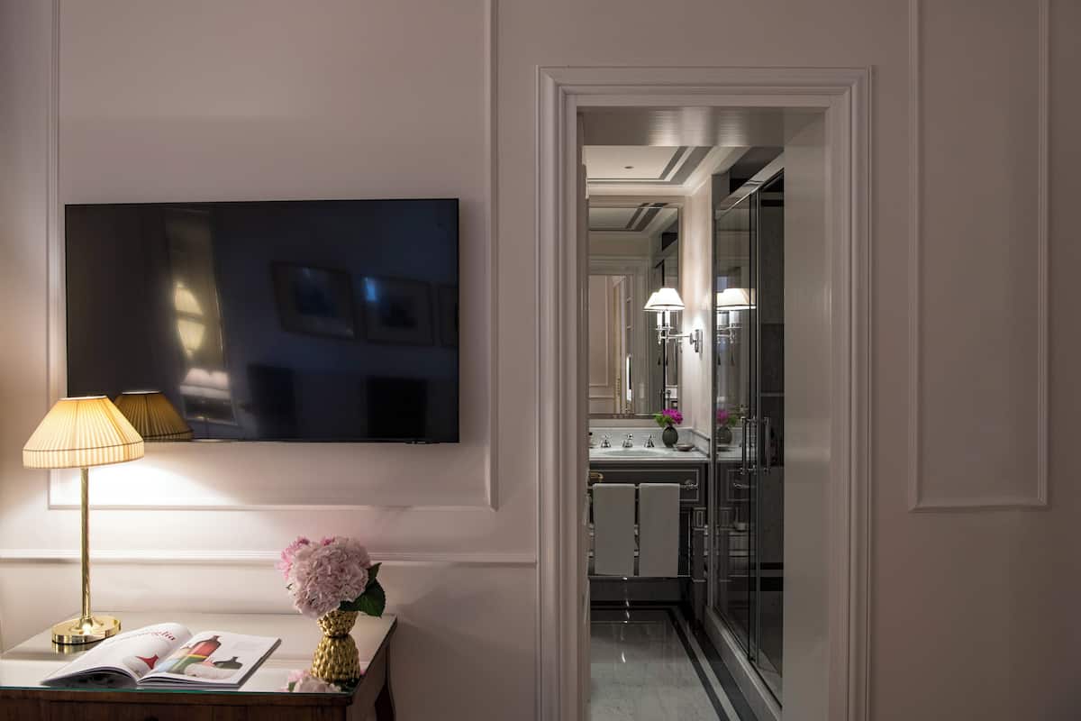 A modern bathroom with a large mirror, a flat-screen TV mounted on the wall, and a vanity with a lamp and flowers.