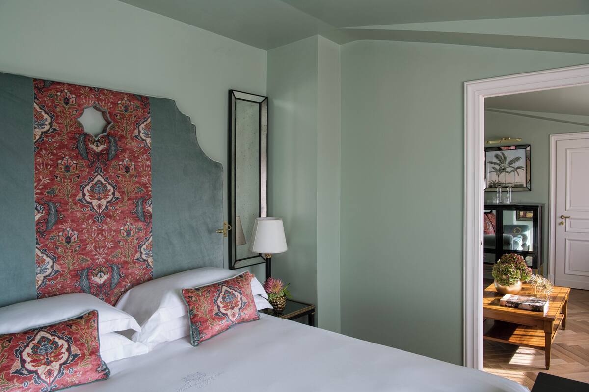 A bedroom with a large bed, a nightstand with a lamp, a decorative wall hanging, and a view into another room.