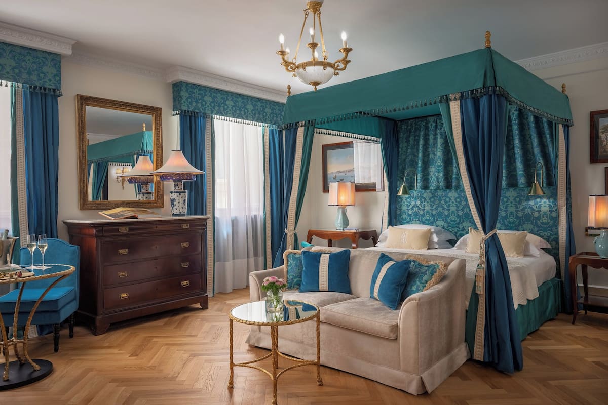A bedroom with a canopy bed, a sitting area with a sofa and armchair, a wooden dresser, and a round coffee table.