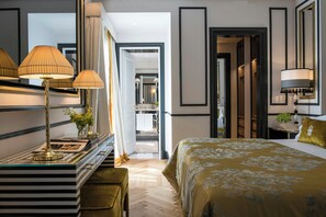 A hotel room with a large bed, a desk with lamps, and a mirror. There is a door leading to a bathroom and a hallway.