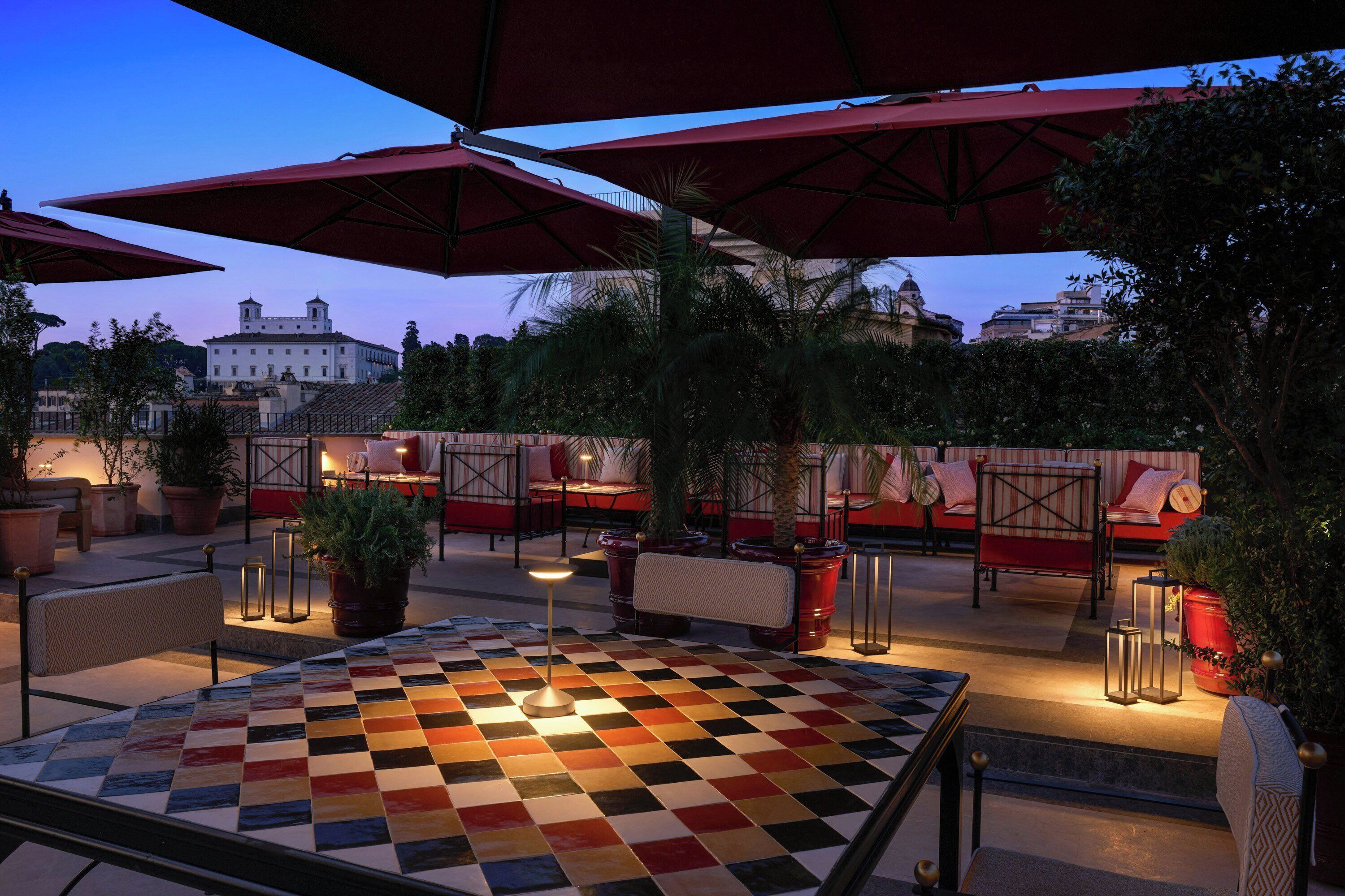 An outdoor seating area with checkered flooring, red umbrellas, and potted plants.