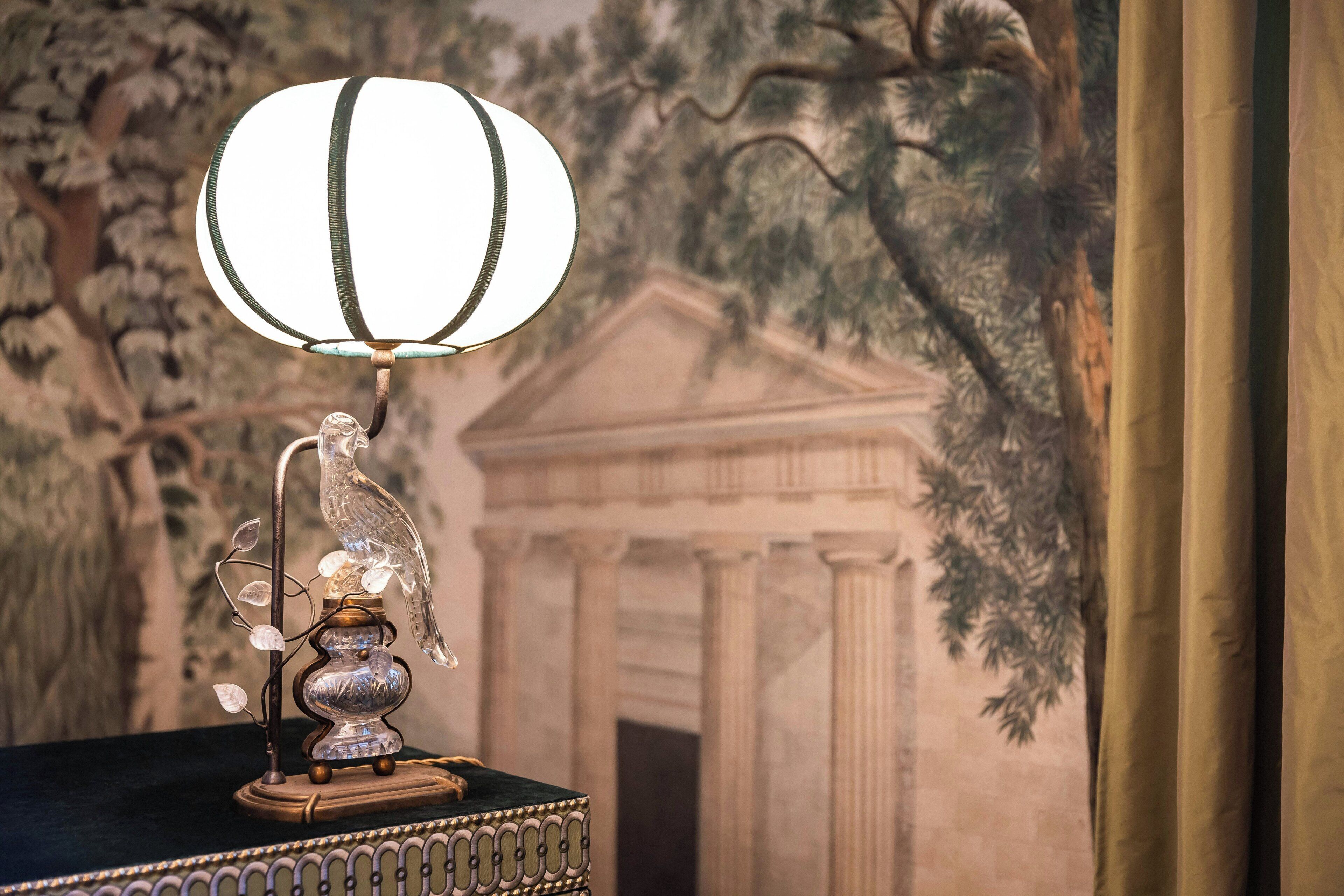 A table lamp with a bird design on a table, set against a mural of a building and trees.