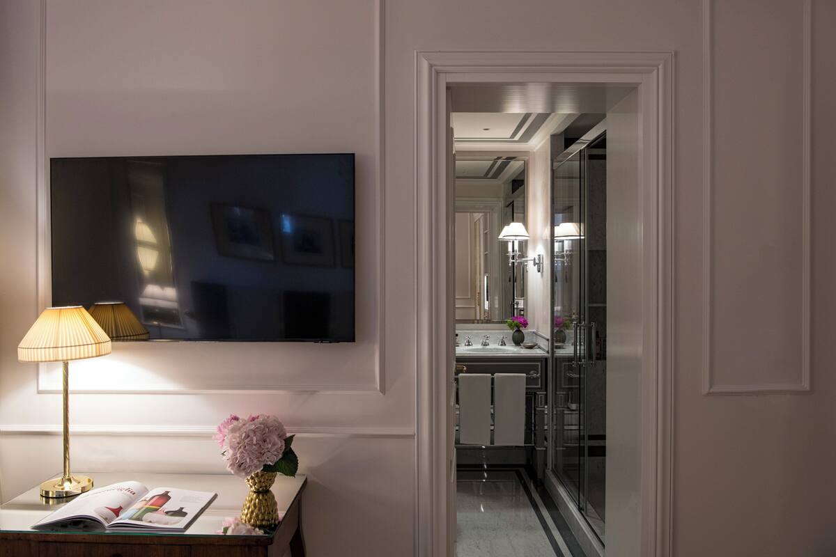 A modern bathroom with a large mirror, a flat-screen TV mounted on the wall, and a vanity with a lamp and flowers.