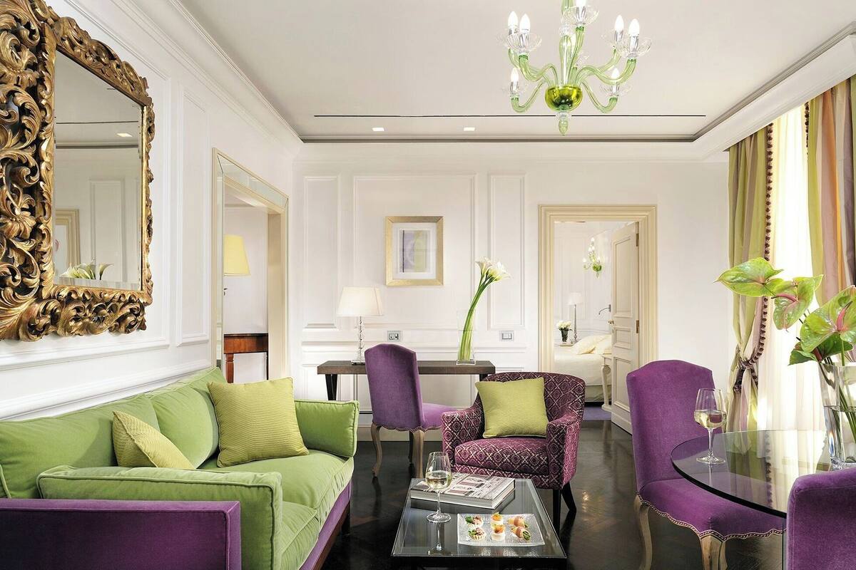 A spacious living room with a green sofa, purple armchairs, a glass coffee table, and a chandelier.