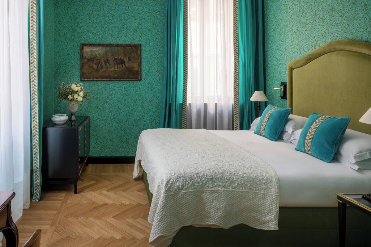 A bedroom with a large bed, teal walls, a painting, a vase with flowers, and a wooden floor.