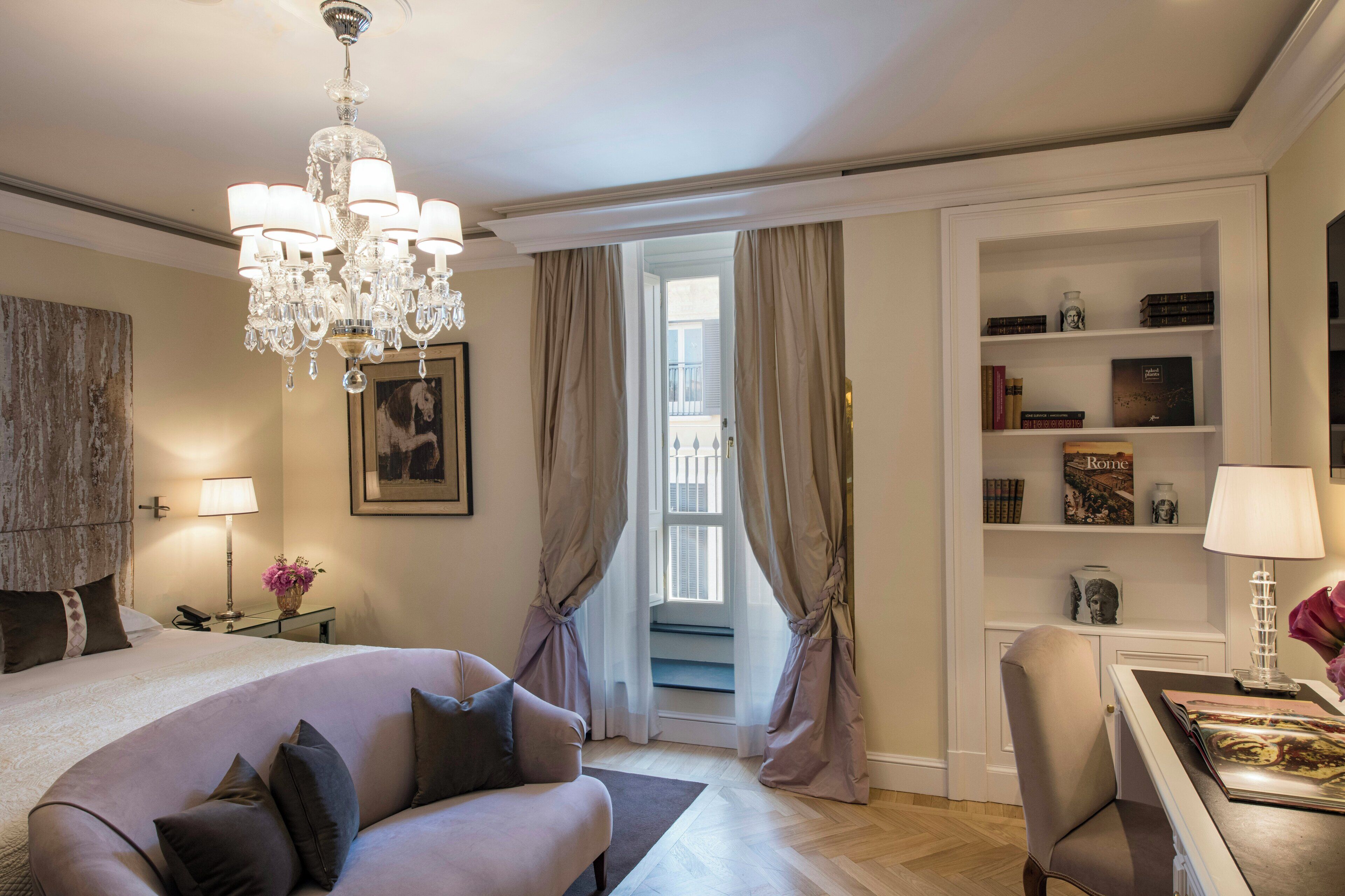 A hotel room with a large bed, a sitting area with a sofa, a chandelier, a bookshelf, and a view of the outside.