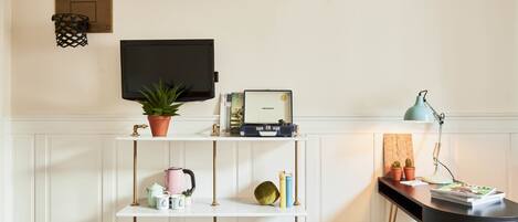 Una habitación con una consola blanca, un escritorio negro con lámpara, televisor montado en la pared y una estantería con objetos decorativos.