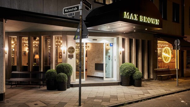 A hotel entrance with a sign reading "Max Brown," a bench, potted plants, and a street sign. - Max Brown Hotel Midtown, part of Sircle Collection (Düsseldorf)