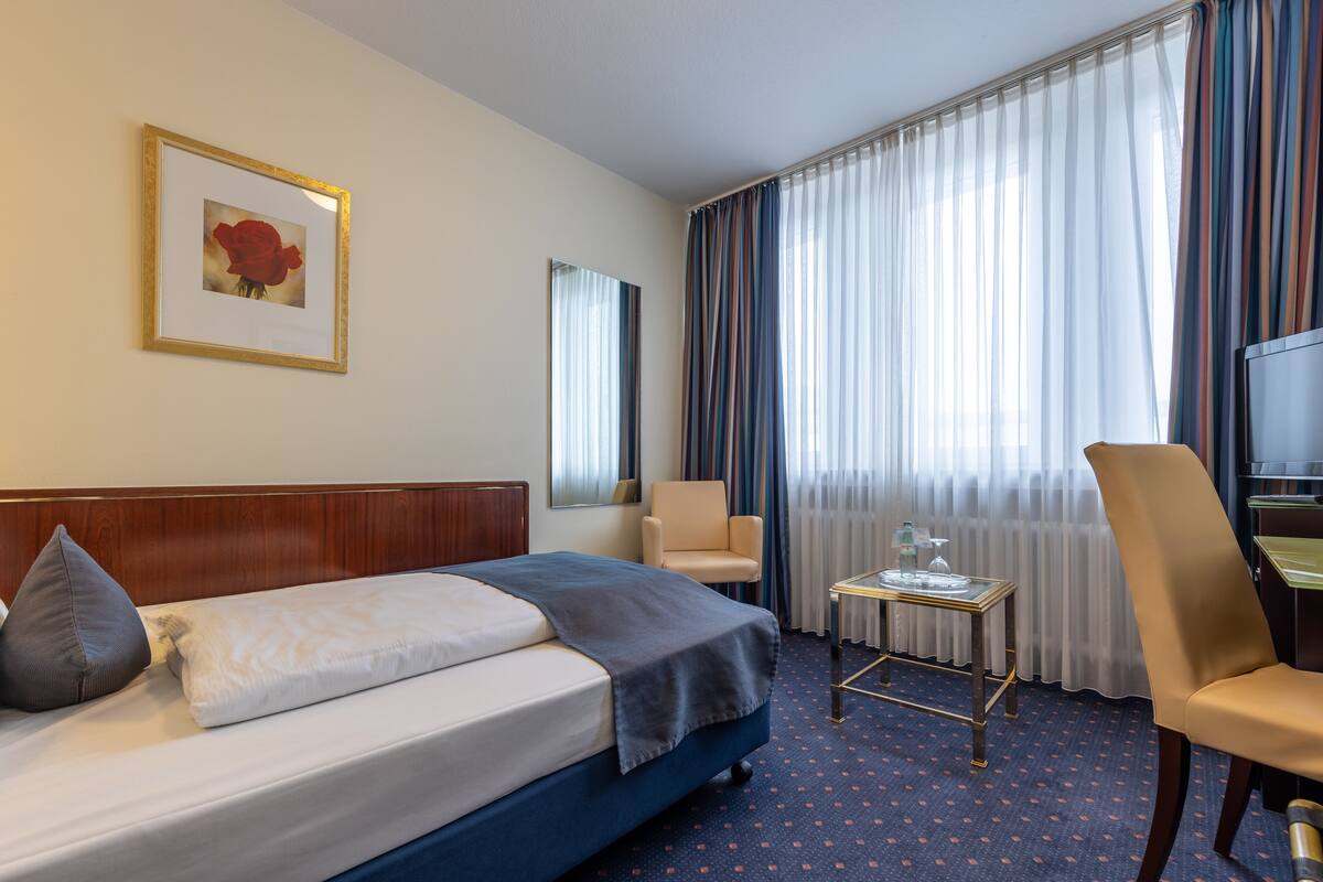 A hotel room with a bed, a chair, a small table, a TV, and a window with curtains.