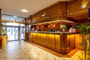 A hotel lobby with a wooden reception desk, clocks, and a large potted plant. - TRIP INN Hotel Uebachs (Düsseldorf)