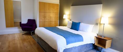 A modern hotel room with a large bed, two bedside lamps, a wooden headboard, a purple chair, and a large mirror.
