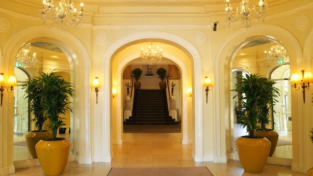 A grand hotel lobby with a marble floor, large potted plants, and a chandelier.