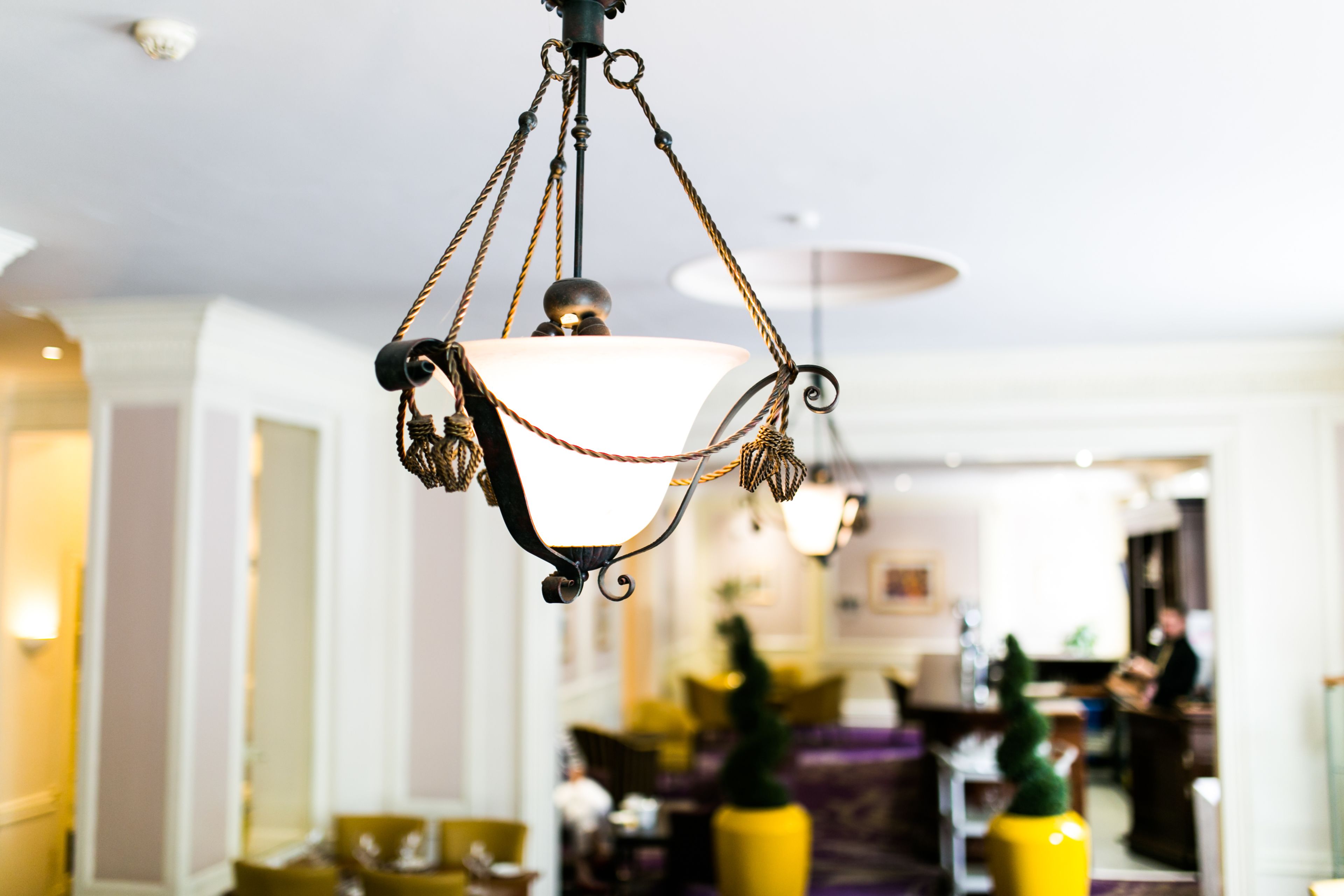 A chandelier with a white shade and a decorative design, hanging in a well-lit indoor space with yellow and purple seating areas.