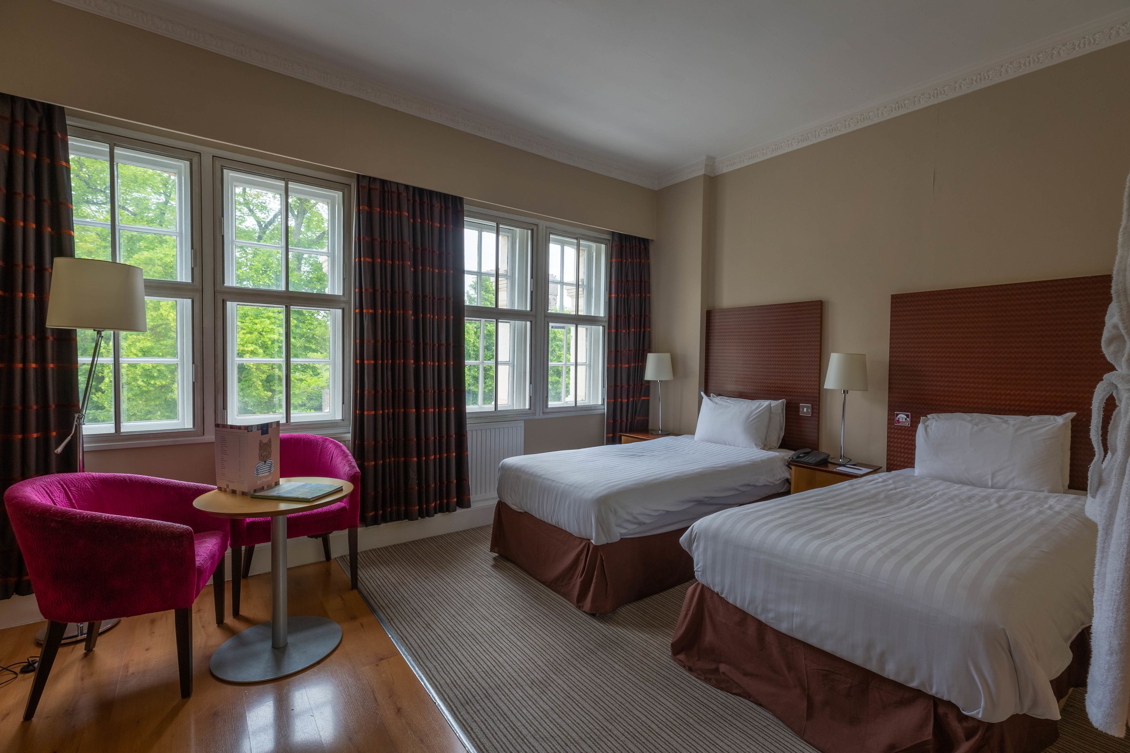A hotel room with two beds, a desk with a chair, and large windows with curtains.