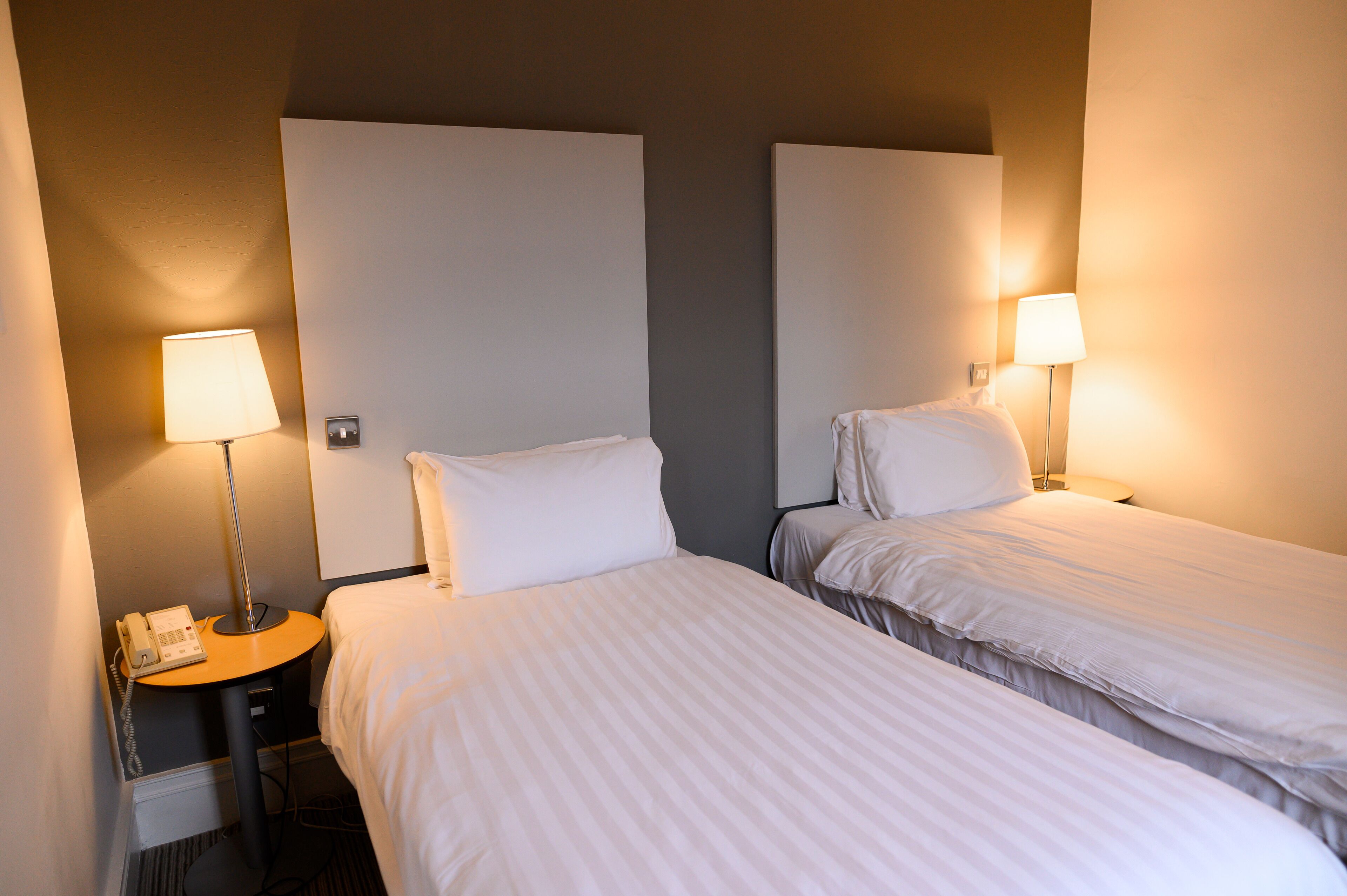 Habitación de hotel con dos camas, cada una con una almohada y una lámpara. Hay una mesita pequeña con un teléfono y una lámpara encima.