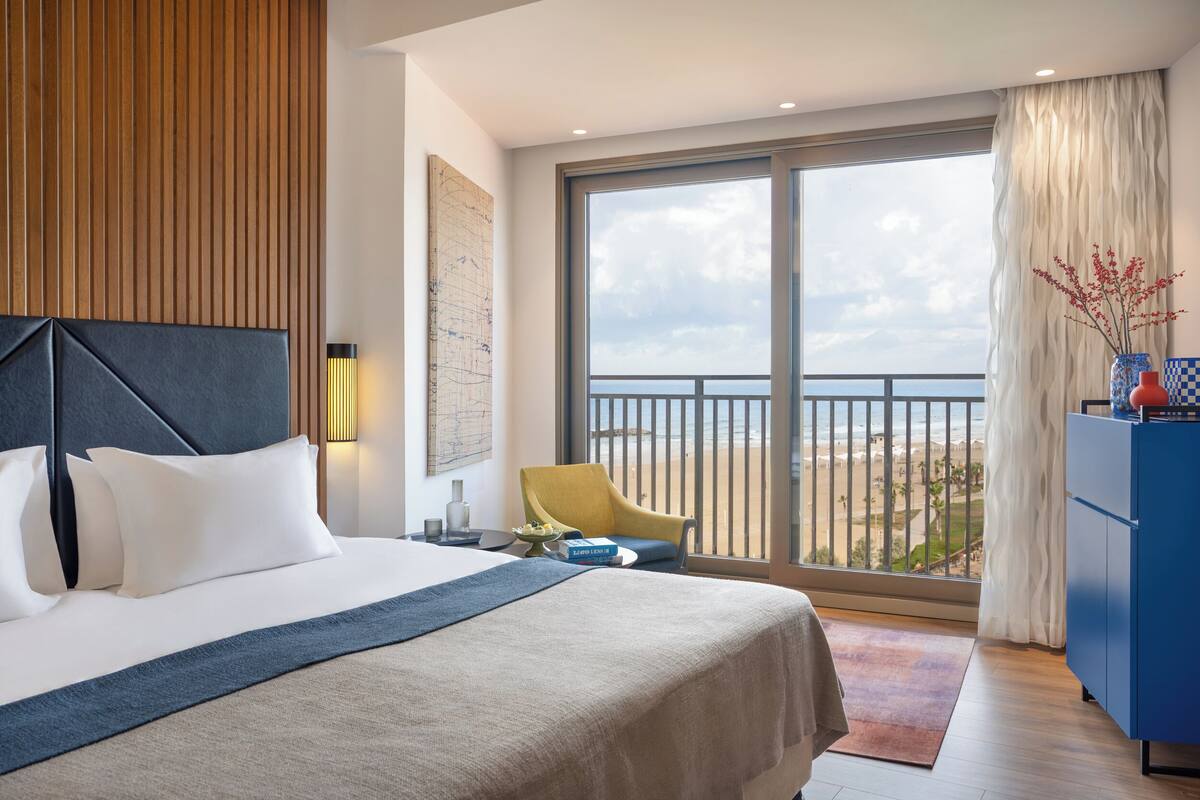 A modern hotel room with a large bed, a view of the beach, a blue cabinet, and a yellow chair.