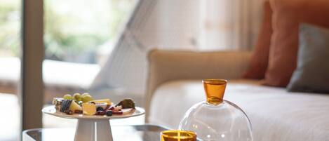 Mesa de vidrio con una copa de vino, un pequeño florero y una bandeja de frutas en una habitación con cama y cortinas.