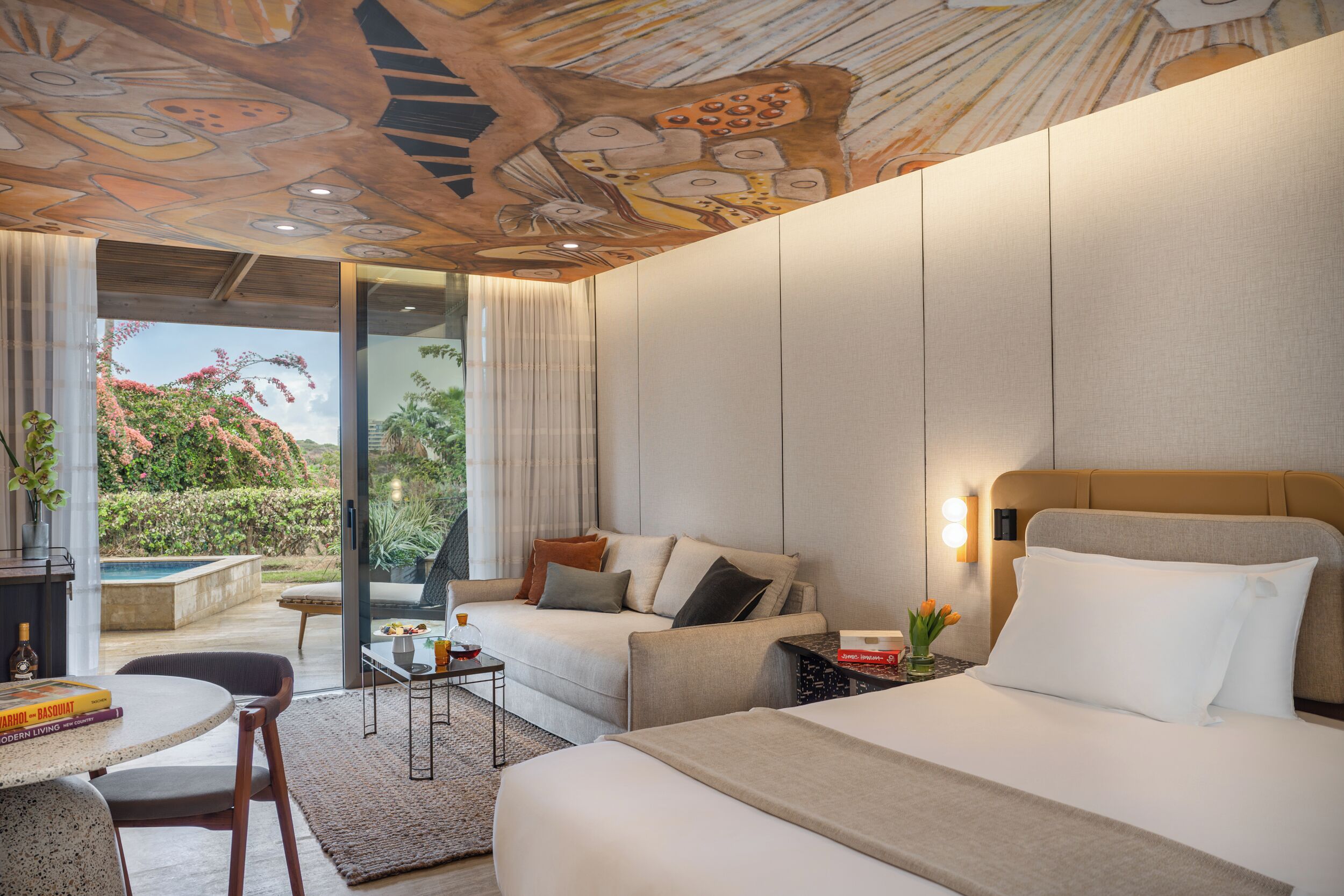 A modern hotel room with a large bed, a sofa, a dining table, and a view of the garden.