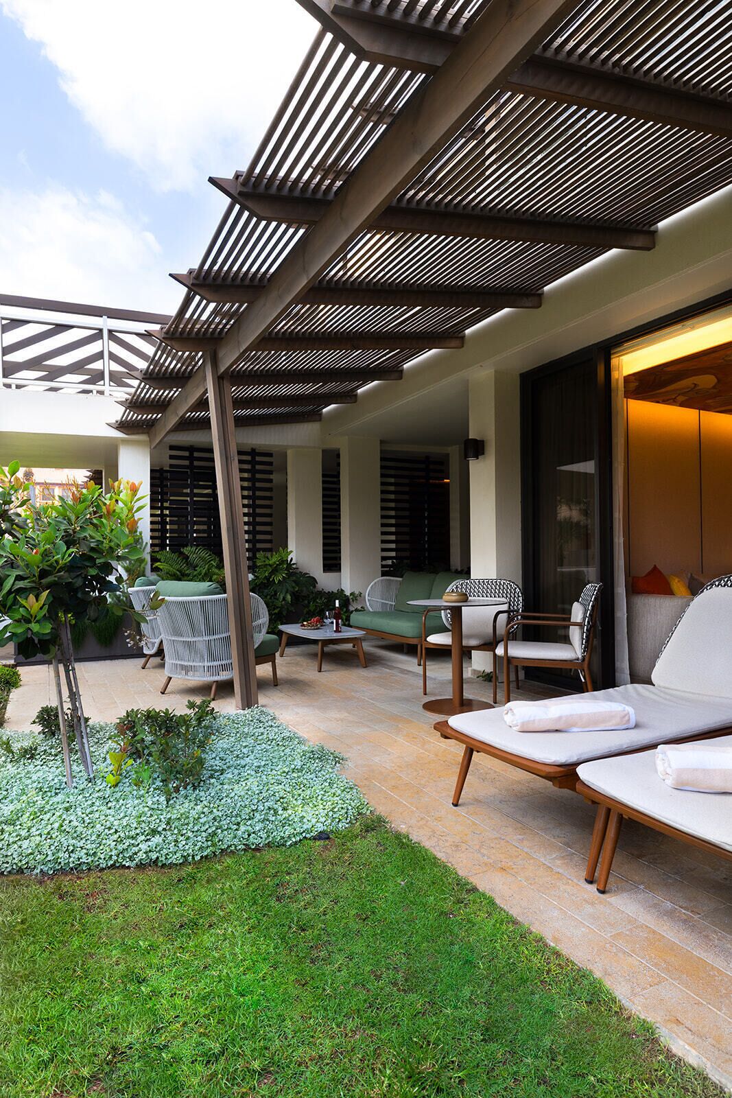 A covered outdoor seating area with lounge chairs and a table, surrounded by greenery and a patterned ceiling.