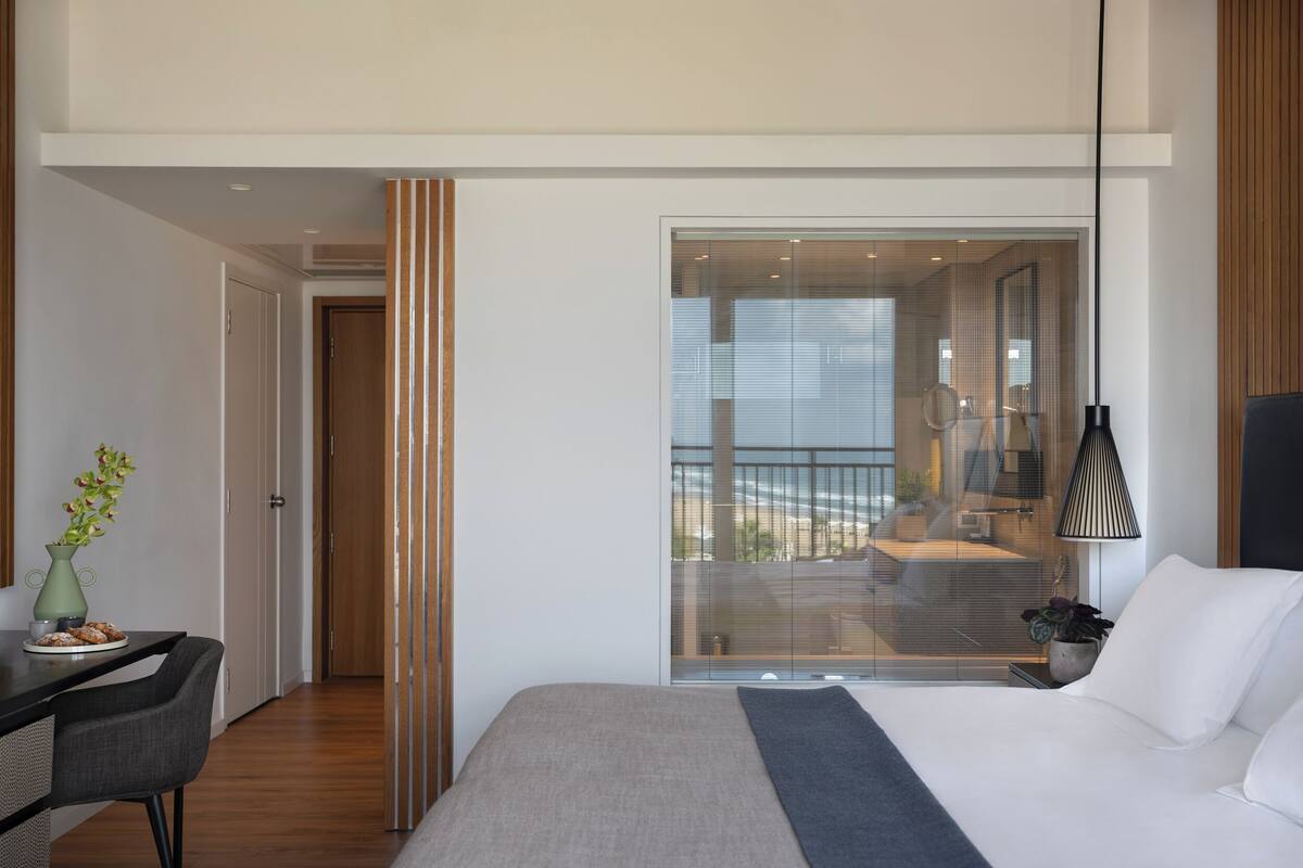 A modern bedroom with a large bed, a desk with a chair, a vase with flowers, and a view of the balcony.