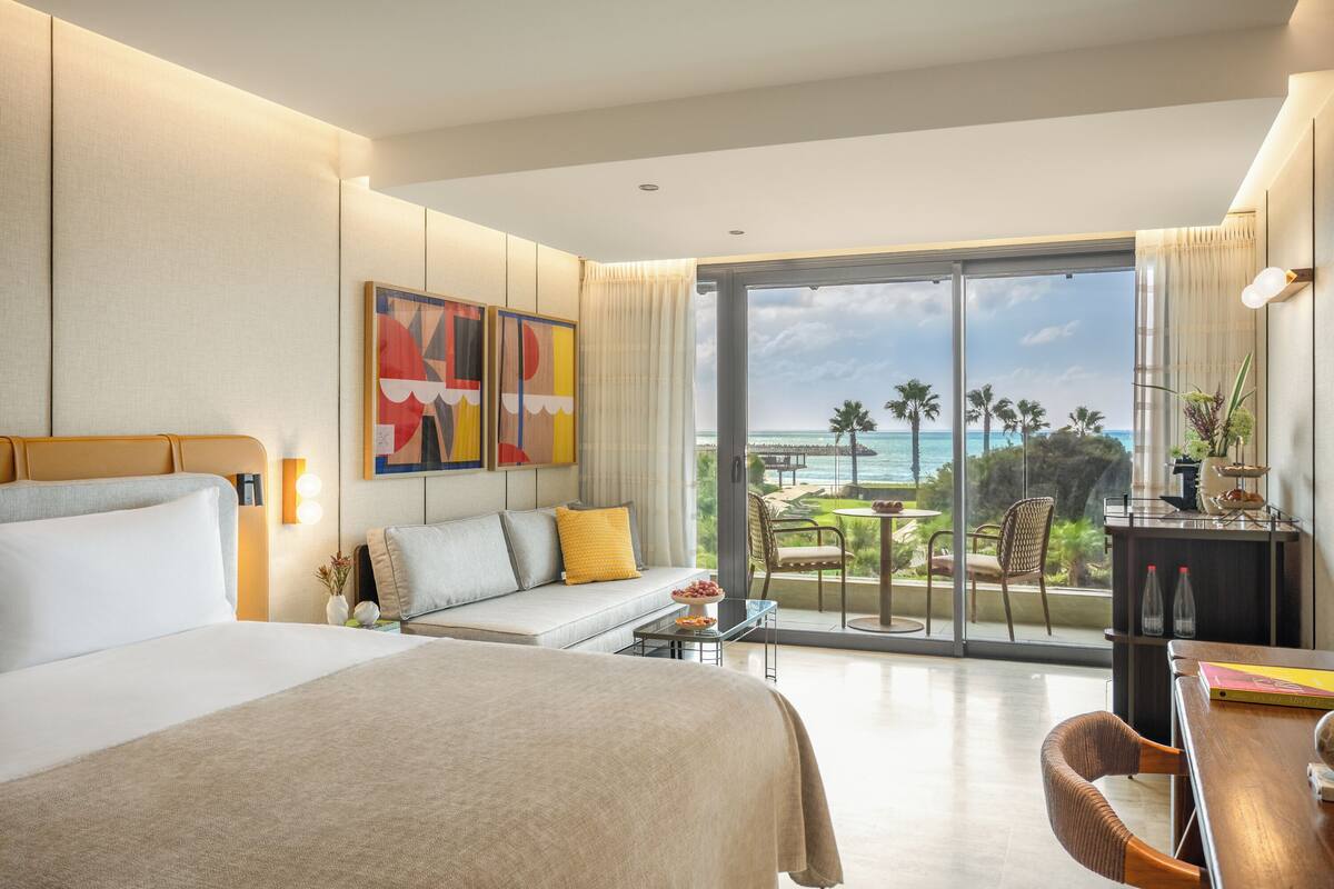 A hotel room with a large bed, a sofa, a dining area, and a balcony with a view of the ocean.