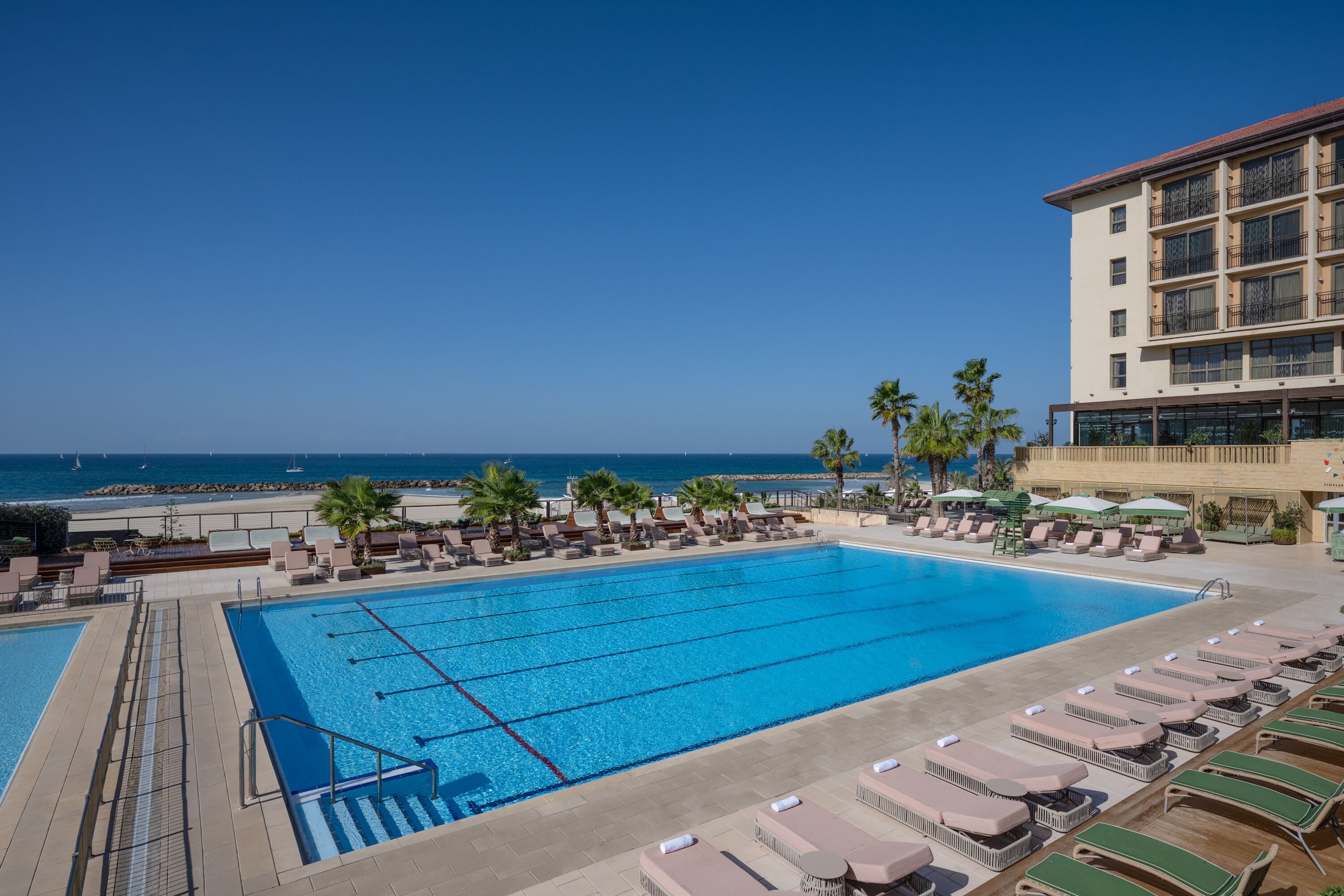 Ein großer Pool mit Liegestühlen und Sonnenschirmen, Blick auf den Strand und ein Gebäude mit Balkonen.