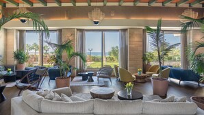 A spacious living area with a wooden ceiling, large windows, and multiple seating options including a sofa, chairs, and a lounge chair. - Dan Accadia Hotel (Herzliya)