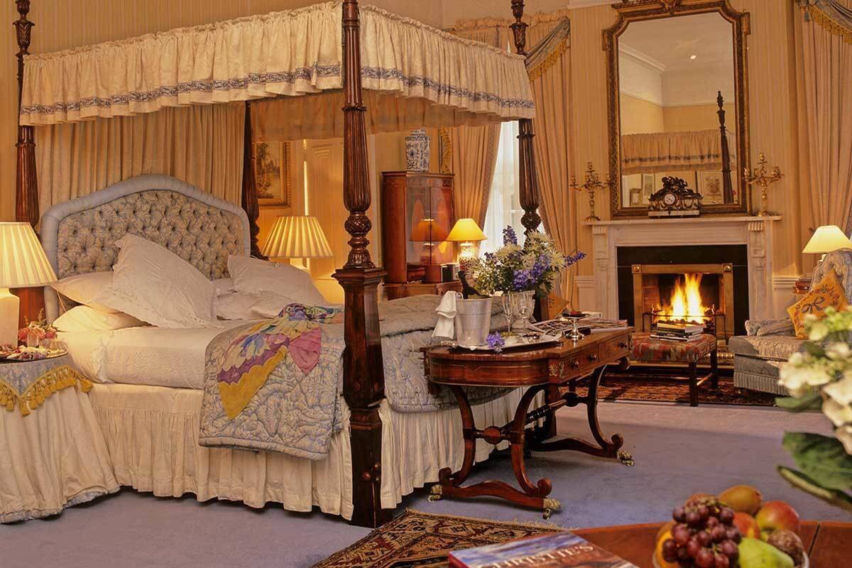 A four-poster canopy bed with a tufted headboard, a bedside table with a lamp, a fireplace with a fire, and a mirror on the wall.