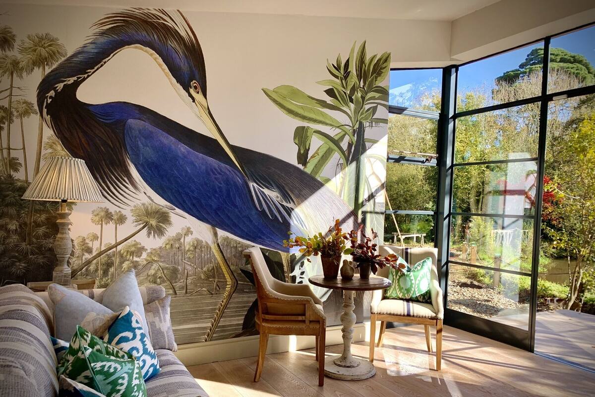 A modern living room with a large mural of a bird, a sofa with patterned cushions, a wooden coffee table with plants, and a view of a garden through large windows.